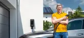 Ein Mann mit einem gelben Poloshirt hält ein blaues E-Ladekabel und steht vor einem E-Auto.