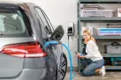 Eine Frau ist in der Hocke in einer Garage und lächelt freundlich. Neben ihr steht ein E-Auto dass an einer Wallbox mit blauem Ladekabel lädt.