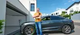 Ein Mann mit einem gelben Poloshirt hält ein blaues E-Ladekabel und steht vor einem E-Auto.