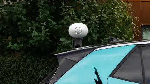 Ein türkises E-Auto steht vor einer grauen Wallbox. Im Hintergrund ist eine dunkelgrüne Hecke zu erkennen.