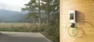 Eine weiße Wallbox mit grünem Ladekabel ist an einer Holzwand montiert. Eine grüne Landschaft ist im Hintergrund zu erkennen.