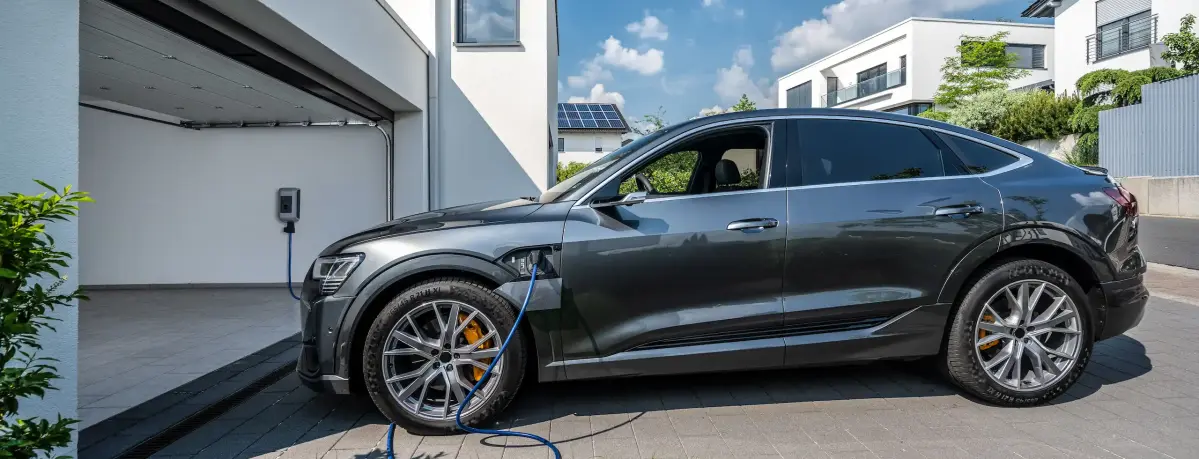 Ein Elektroauto lädt an einer Ladestation in einer modernen Wohngegend auf. Im Hintergrund sind ein offenes Garagentor, ein Solarpanel und andere Gebäude zu sehen.