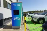 Eine Ladestation mit der Aufschrift „Löhr Gruppe eFix“ lädt Elektroautos auf einem Parkplatz. Im Hintergrund sind grüne Parkplätze und mehrere geparkte Autos zu sehen.