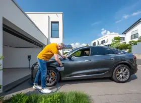 Ein Mann mit einem gelben Poloshirt hält ein blaues E-Ladekabel und lädt ein E-Auto.