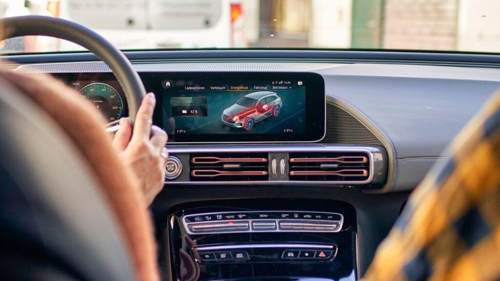 Eine Person sitzt am Steuer eines Autos und eine Person sitzt auf dem Beifahrersitzt. Das Display des Autos ist in der Mitte des Bildes