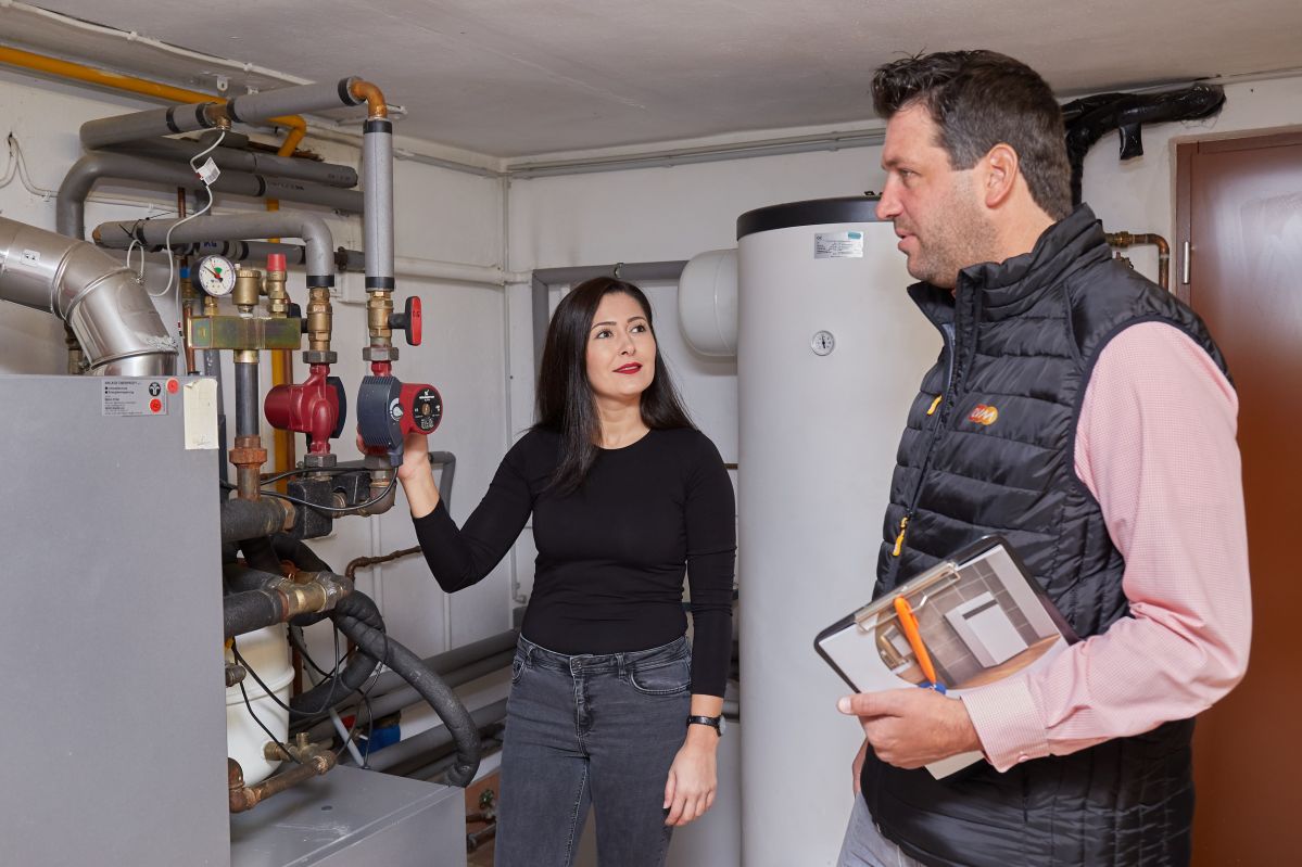 Ein Mann mit einer schwarzen evm-Weste steht in einem Heizungskelller mt einer Frau.