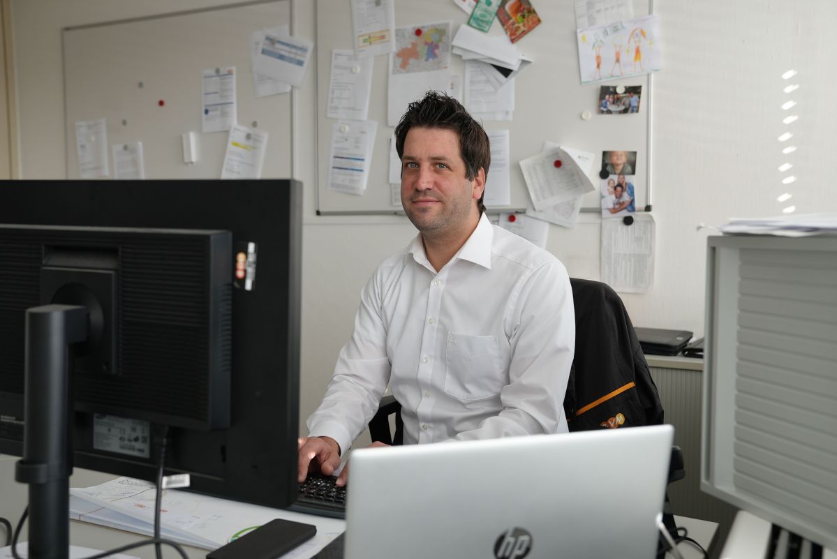 Ein Mann sitzt an einem Schreibtisch und arbeitet am Computer. Der Raum ist mit einem Whiteboard voller Notizen im Hintergrund ausgestattet. Ordner und Papiere liegen herum.