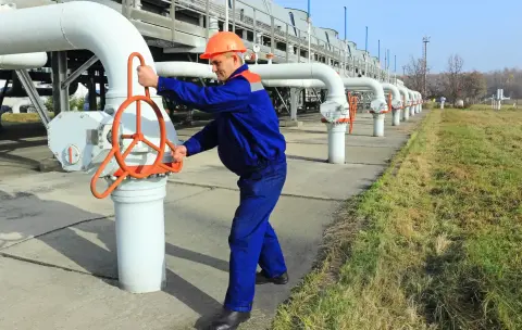 Ein Mann mit blauer Arbeitskleidung und einem Schutzhelm dreht ein orangenes Gasrohr zu.