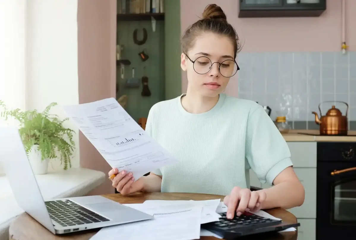 Eine junge Frau mit Brille sitzt an einem Küchentisch und tippt auf einem Taschenrechner. Sie hält Dokumente in der Hand und vor ihr steht ein Laptop.