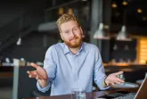 Ein Mann mit einem blauen Hemd sitzt an einem Tisch und macht eine Geste die "keine Ahnung" bedeuten soll. Vor ihm steht ein Laptop