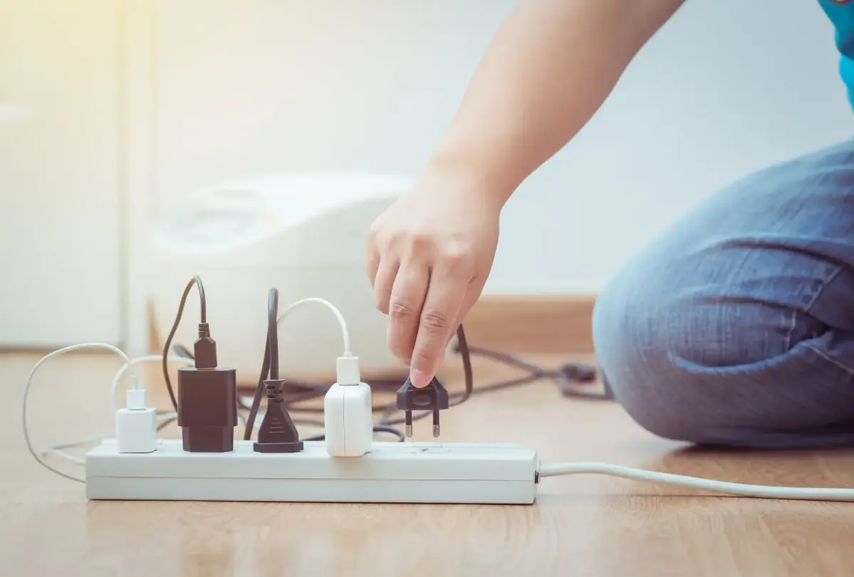 Eine Hand steckt einen Stecker in eine weiße Mehrfachsteckdose mit mehreren Kabeln; im Hintergrund ist ein weißes Haushaltsgerät auf Holzboden zu sehen.