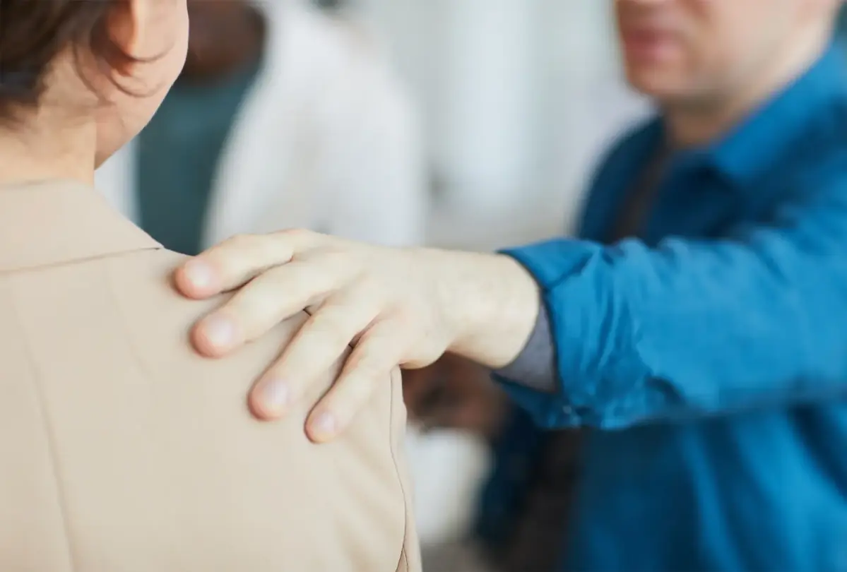 Eine Hand liegt beruhigend auf der Schulter einer Person in hellbrauner Kleidung. Im Hintergrund sind unscharf zwei weitere Menschen erkennbar.