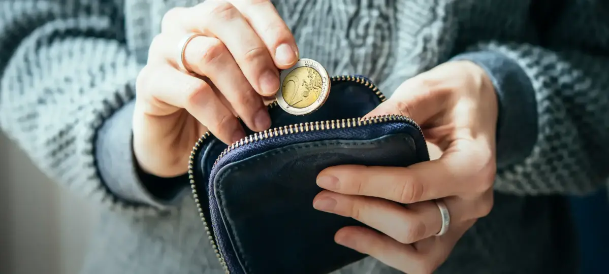 Eine Person hält eine blaue Geldbörse und steckt eine Euro-Münze hinein. Die Person trägt einen grauen Pullover, und die Umgebung wirkt alltäglich und ruhig.