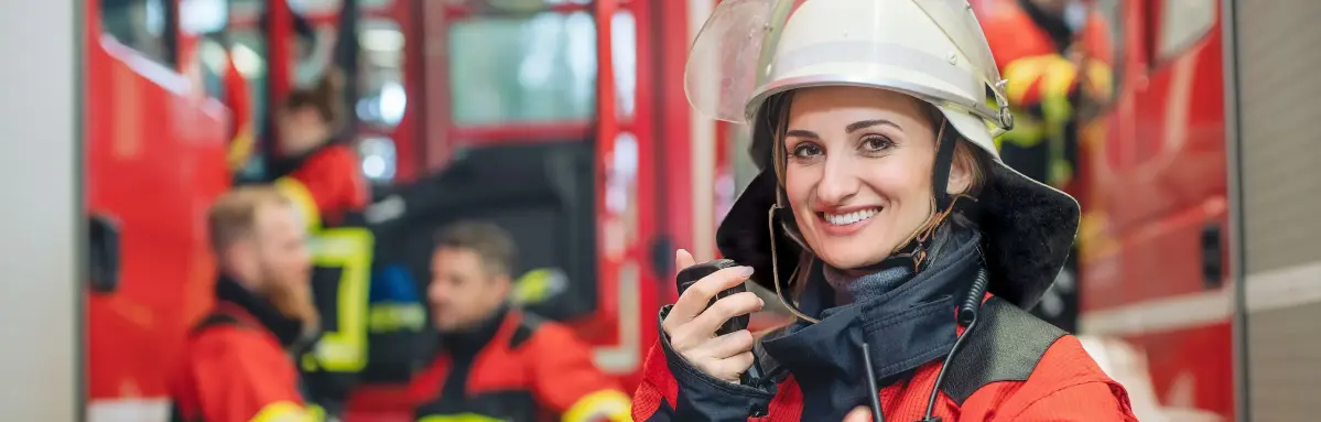 Energieschub evm Feuerwehr Eine Feuerwehrfrau lächelt und hält ein Funkgerät, umgeben von roten Feuerwehrautos und anderen Feuerwehrleuten in Ausrüstung.