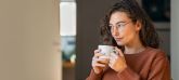 Eine Person mit lockigem Haar und Brille hält eine Tasse und schaut nachdenklich zur Seite. Sie trägt einen orangefarbenen Pullover, im Hintergrund ein Innenraum mit weichen Lichtverhältnissen.