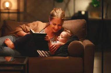 Eine Frau liegt mit ihrer Tochter auf einem Sofa und liest aus einem Buch vor. Auf dem Sofatisch steht ein Glas mit einer Lichterkette.