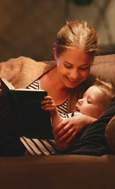 Eine Frau liegt mit ihrer Tochter auf einem Sofa und liest aus einem Buch vor. Auf dem Sofatisch steht ein Glas mit einer Lichterkette.