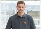 Engel Sascha_307 1 Ein Mann steht ruhig, trägt ein graues Poloshirt mit einem roten und gelben Logo "evm". Im Hintergrund bietet ein Fensterblick eine verschwommene Stadtlandschaft.