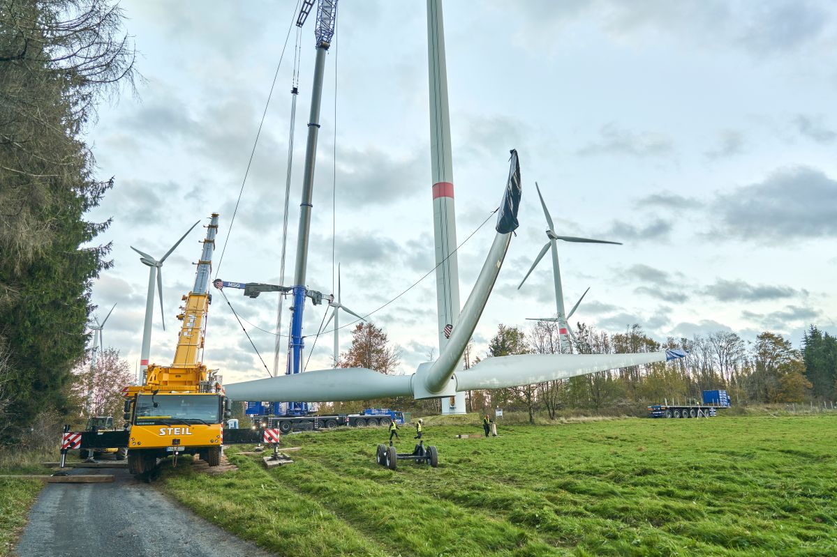 evm Windenergie Ein Kran hebt ein Windrad-Blatt, während Techniker auf einer Wiese arbeiten; im Hintergrund weitere Windräder und Bäume.