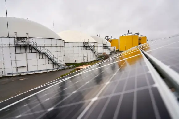 Im Vordergrund ist eine dunkle PV-Anlage zu erkennen. Links i Hintergrund befinden sich große weiße und gelbe Kessel.