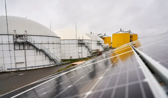 Im Vordergrund ist eine dunkle PV-Anlage zu erkennen. Links i Hintergrund befinden sich große weiße und gelbe Kessel.