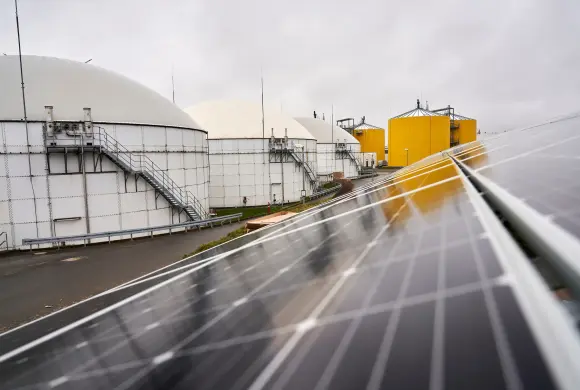 Im Vordergrund ist eine dunkle PV-Anlage zu erkennen. Links i Hintergrund befinden sich große weiße und gelbe Kessel.