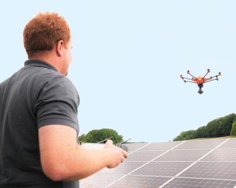Ein Mann mit einem dunkelgrauen Poloshirt bedient eine Drohne und fliegt diese über Module in einem Solarpark.