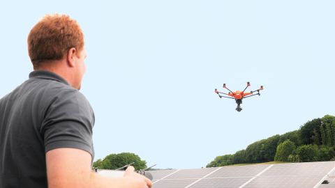 Ein Mann mit einem dunkelgrauen Poloshirt bedient eine Drohne und fliegt diese über Module in einem Solarpark.