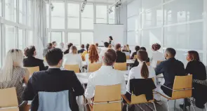 Indian businesswoman leading the seminar Eine Gruppe von Menschen sitzt auf Stühlen und hört einer Person zu, die vorne in einem hellen Raum vor einem Projektionsbildschirm spricht.
