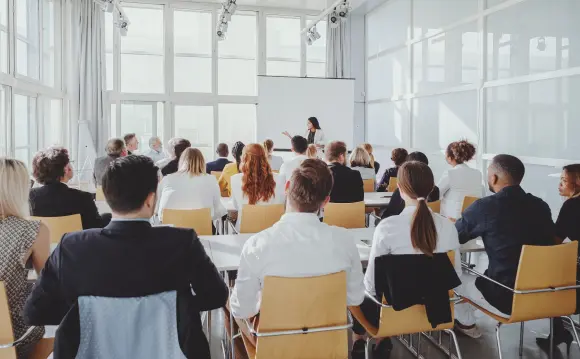 Eine Gruppe von Menschen sitzt auf Stühlen und hört einer Person zu, die vorne in einem hellen Raum vor einem Projektionsbildschirm spricht.