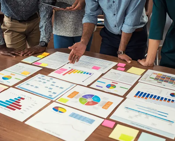 Mehrere Personen sind über einen großen Tisch gebeugt. Auf dem Tisch liegen viele Papiere mit bunten Grafiken und Statistiken.