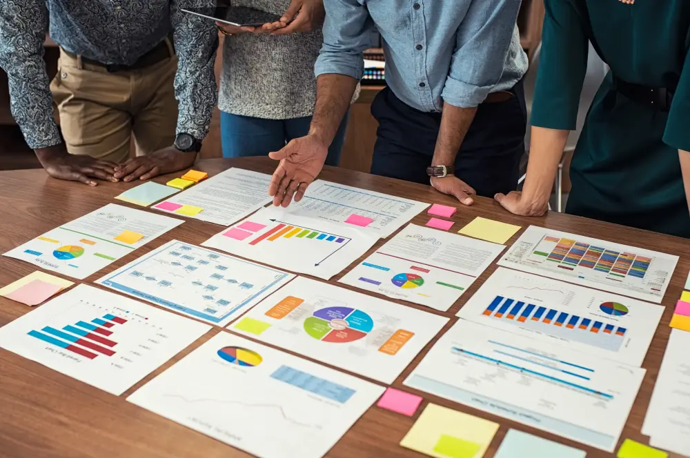Business casual team working on graphs Mehrere Personen sind über einen großen Tisch gebeugt. Auf dem Tisch liegen viele Papiere mit bunten Grafiken und Statistiken.