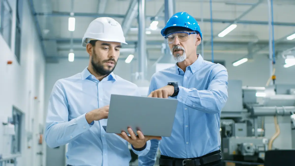 782842951 Zwei professionell gekleidete Männer mit Schutzhelmen stehen in einer Fabrik und schauen auf einen Laptop.