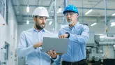 782842951 Zwei professionell gekleidete Männer mit Schutzhelmen stehen in einer Fabrik und schauen auf einen Laptop.