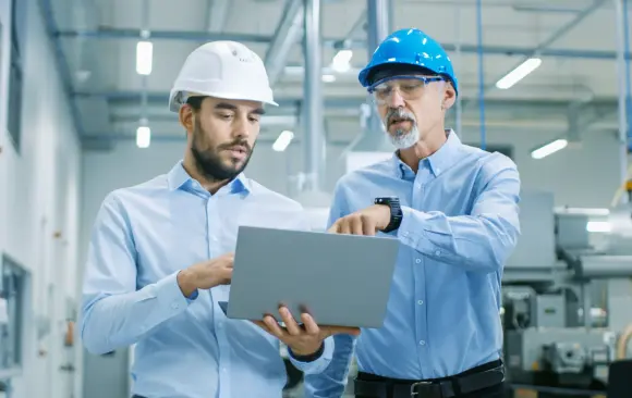 Zwei professionell gekleidete Männer mit Schutzhelmen stehen in einer Fabrik und schauen auf einen Laptop.