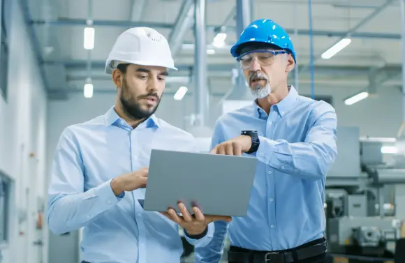 782842951 Zwei professionell gekleidete Männer mit Schutzhelmen stehen in einer Fabrik und schauen auf einen Laptop.