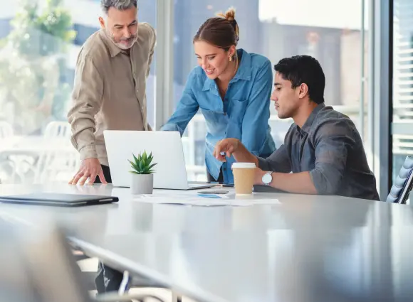 Zwei professionell gekleidete Männer und eine Frau sitzen an einem Besprechungstisch. Sie schauen auf einen Laptop und diskutieren.