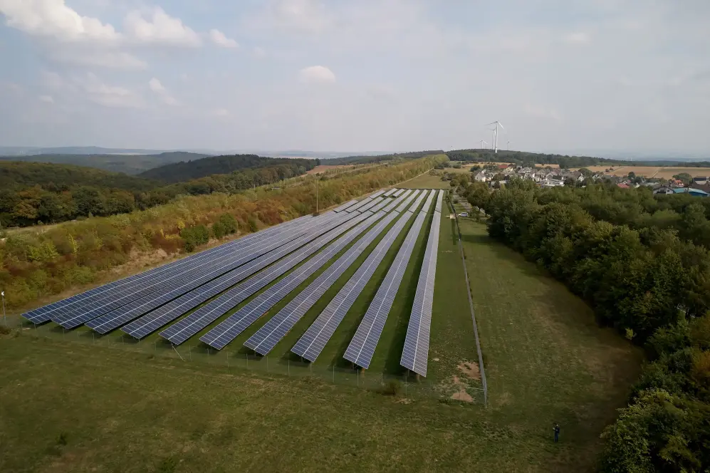 Himmelblick auf einen Solarpark in einer begrünten Umgebung und blauem Himmel mit weißen Wolken.