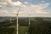 Mehrere Windräder drehen sich in einer grünen Landschaft, umgeben von Wäldern und unter einem blauen Himmel mit weißen Wolken.