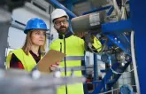 Eine Frau und ein Mann mit gelber Schutzkleidung und Schutzhelmen stehen in einer Fabrik und führen eine Überprüfung durch. Die Frau hält ein Cliipboard in der Hand.