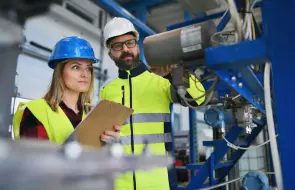 Eine Frau und ein Mann mit gelber Schutzkleidung und Schutzhelmen stehen in einer Fabrik und führen eine Überprüfung durch. Die Frau hält ein Cliipboard in der Hand.