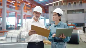 2286642833 Ein Mann mit Schutzhelm und einem unterhält sich mit einer Frau die einen Computer hält in einer Fabrikumgebung. Beide tragen Arbeitskleidung.