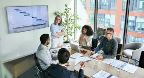 Eine Gruppe von fünf Personen diskutiert um einen Tisch. Präsentationen und Notizen liegen aus. Ein Bildschirm zeigt ein Diagramm. Der Raum hat große Fenster mit Blick auf ein Gebäude.