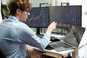 Young investor in stock trader analyzing on dynamic exchange screen. Gusher. Ein Mann zeigt mit einem Stift auf zwei Monitore mit Finanzdiagrammen, während er an einem Schreibtisch sitzt. Er trägt ein blaues Hemd und eine Brille. Im Hintergrund sind Pflanzen.