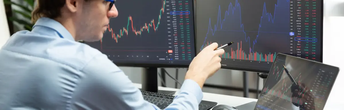Ein Mann zeigt mit einem Stift auf zwei Monitore mit Finanzdiagrammen, während er an einem Schreibtisch sitzt. Er trägt ein blaues Hemd und eine Brille. Im Hintergrund sind Pflanzen.