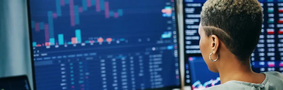 2364201097 Eine Frau arbeitet am Computer, analysiert Finanzdaten auf mehreren Bildschirmen. Der Schreibtisch ist mit Notizbuch, Stift und Tastatur ausgestattet, ein Bürohintergrund ist sichtbar.