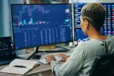 2364201097 Eine Frau arbeitet am Computer, analysiert Finanzdaten auf mehreren Bildschirmen. Der Schreibtisch ist mit Notizbuch, Stift und Tastatur ausgestattet, ein Bürohintergrund ist sichtbar.
