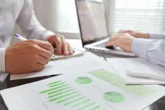 279358256 Zwei Personen sitzen an einem Besprechungstisch. Vor ihnen steht ein Computer und es liegen Unterlagen mit Diagrammen und Statistiken auf dem Tisch.