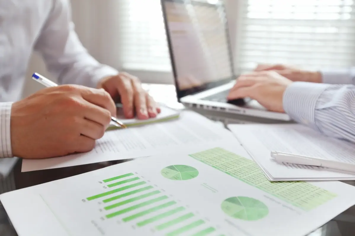 Zwei Personen sitzen an einem Besprechungstisch. Vor ihnen steht ein Computer und es liegen Unterlagen mit Diagrammen und Statistiken auf dem Tisch.