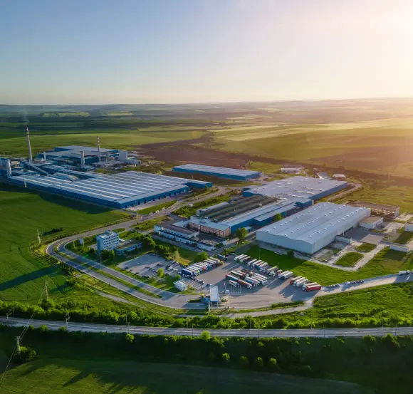 Ein großes Fabrikgelände liegt inmitten einer weiten, grünen Landschaft unter einem sonnigen Himmel. Mehrere Gebäude und geparkte Transportfahrzeuge sind sichtbar, umgeben von Feldern und Straßen.